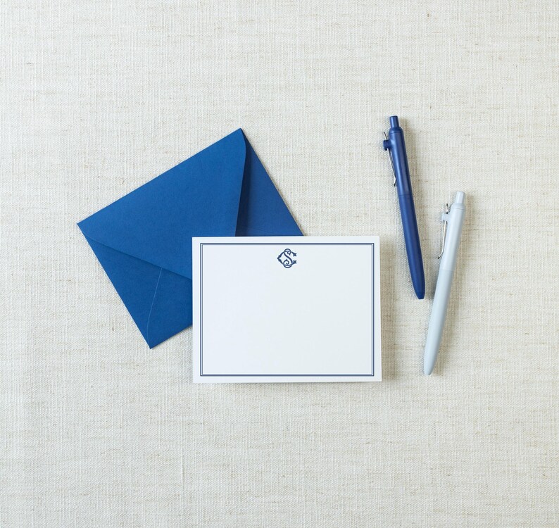 May include: A white rectangular note card with a dark blue border and a central monogram, accompanied by a matching dark blue envelope, a dark blue retractable ballpoint pen, and a light gray retractable ballpoint pen.