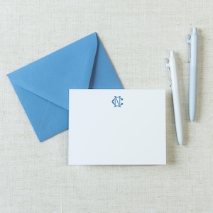 May include: A white rectangular note card featuring a blue monogram with the letters C and N, accompanied by a matching blue envelope and two light blue ballpoint pens. The stationery set is arranged neatly to display the centered blue emblem.