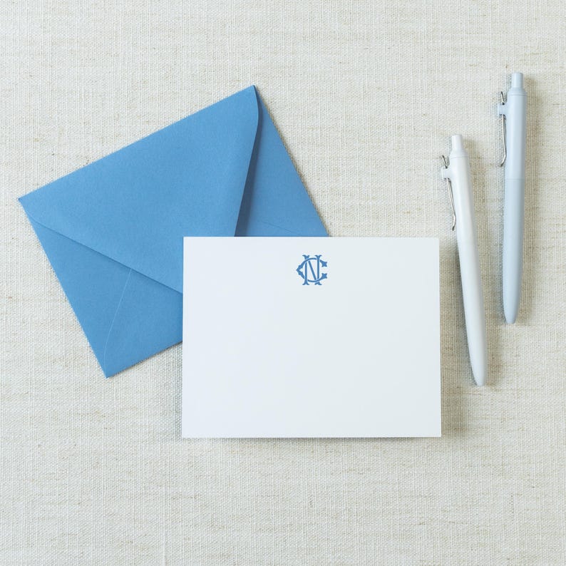 May include: A white rectangular note card featuring a blue monogram with the letters C and N, accompanied by a matching blue envelope and two light blue ballpoint pens. The stationery set is arranged neatly to display the centered blue emblem.
