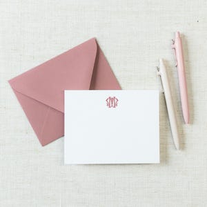 May include: A white rectangular note card featuring a centered pink monogram, accompanied by a matching pink envelope and two pens, one in a light pink shade and the other in a slightly darker pink, arranged on a textured off-white surface.