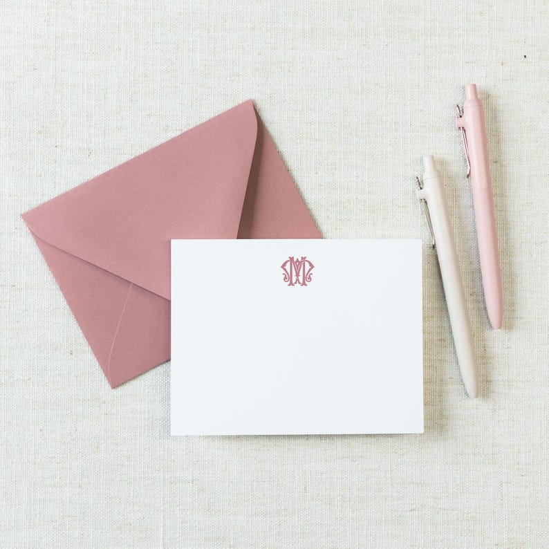 May include: A white rectangular note card featuring a centered pink monogram, accompanied by a matching pink envelope and two pens, one in a light pink shade and the other in a slightly darker pink, arranged on a textured off-white surface.