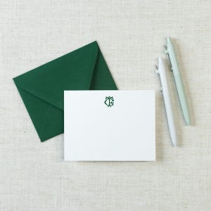 May include: A white rectangular note card featuring a dark green monogram of the letters T and G, accompanied by a dark green envelope and two light green pens. The stationery set is arranged neatly to showcase the centered green emblem.