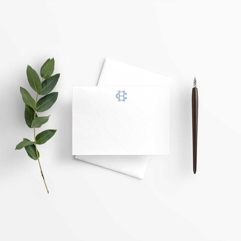 May include: A white rectangular note card featuring a centered light blue monogram with the letter H, accompanied by a white envelope, a dark brown fountain pen, and a green eucalyptus branch with several leaves arranged on the side.