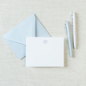 May include: A light blue envelope and a white rectangular note card featuring a centered gray decorative monogram emblem. Two pens, one light blue and one white, are positioned to the right of the stationery set on a textured surface.
