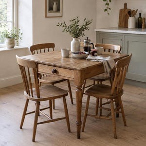 May include: A square wooden dining table with a single drawer featuring a round knob, accompanied by four matching wooden spindle-back chairs. The table surface holds a white ceramic vase with greenery, a bowl of berries, a French press, and mugs.