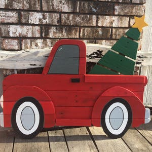 May include: A red wooden pickup truck with a green wooden Christmas tree in the bed. The truck has white tires and a gray cab. The truck is painted with a rustic, distressed look.