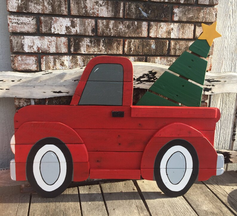May include: A red wooden pickup truck with a green wooden Christmas tree in the bed. The truck has white tires and a gray cab. The truck is painted with a rustic, distressed look.