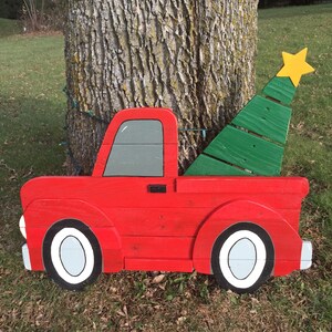 May include: A red wooden pickup truck with a green wooden Christmas tree in the bed. The truck has white wheels and a gray window. The tree has a yellow star on top.