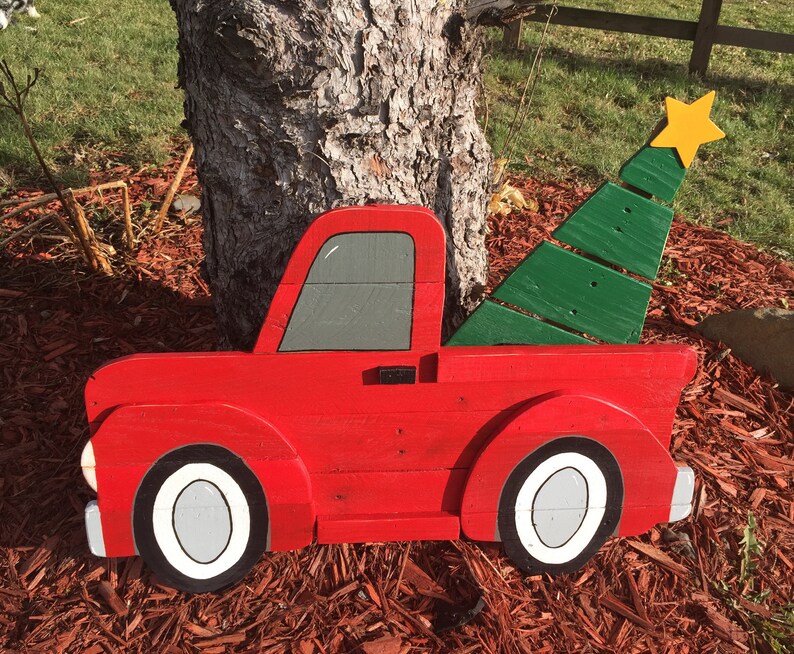 May include: A red wooden pickup truck with a green wooden Christmas tree in the bed. The truck has white tires and a gray window. The tree has a yellow star on top.