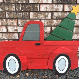 May include: A red wooden pickup truck with a green wooden Christmas tree in the bed. The truck has white wheels and a gray cab. The truck is painted in a rustic style.