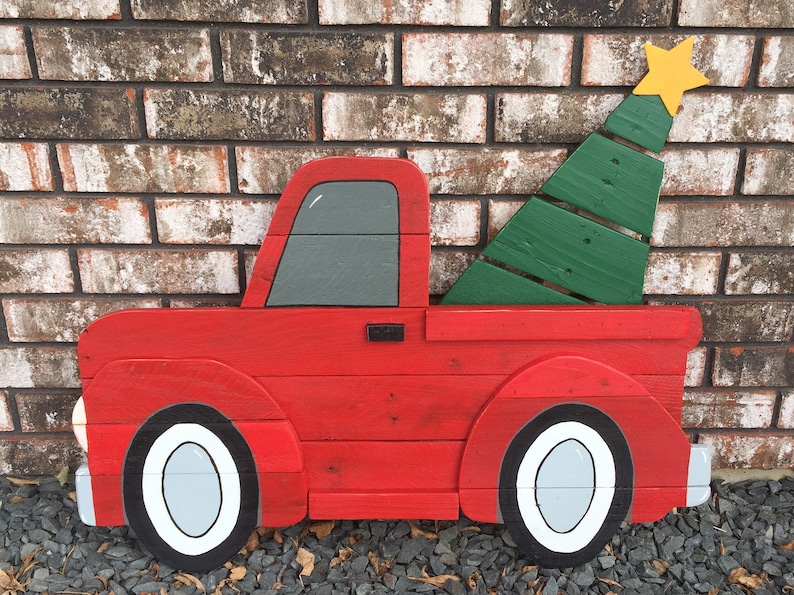 May include: A red wooden pickup truck with a green wooden Christmas tree in the bed. The truck has white wheels and a gray cab. The truck is painted in a rustic style.