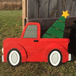 May include: A red wooden toy pickup truck with a green wooden Christmas tree in the bed. The truck has white tires and a gray window. The tree has a yellow star on top.
