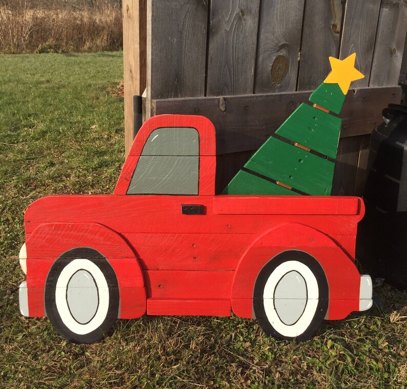May include: A red wooden toy pickup truck with a green wooden Christmas tree in the bed. The truck has white tires and a gray window. The tree has a yellow star on top.