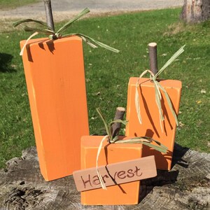 May include: Three orange wooden pumpkins with green stems and twine accents. The smallest pumpkin has a brown wooden sign that says "Harvest".