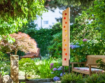 May include: A wooden outdoor light with coloured glass squares set in a garden. The light is supported by black metal legs. The garden features a wooden bench, trees, and colourful flowers. The light's height is approximately 180 cm.