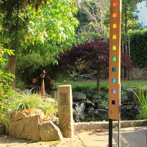 Könnte beinhalten: Ein Gartenpfahl aus Holz mit einem Regenbogen aus farbigem Glasquadraten. Der Pfahl steht in einem Garten mit einem kleinen Teich und einer Steinsäule.
