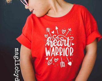 May include: A red t-shirt with the words "heart warrior" in white script and decorative arrows and hearts. The shirt is worn by a young child wearing pink sunglasses.