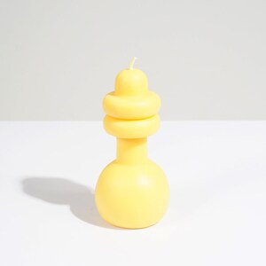May include: A yellow candle with a unique, layered design. The candle is shaped like a vase with a rounded base and a layered top.