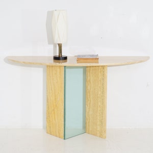 May include: A half-round travertine console table with a glass panel in the center of the base. The table has a light beige color and is topped with a white lamp and a book.