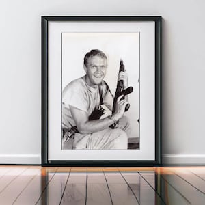 Könnte beinhalten: Schwarz-Weiß-Druck in einem schwarzen Rahmen, der einen lächelnden Mann mit einem Gewehr zeigt. Das Foto ist in einem schwarzen Rahmen vor einer weißen Wand. Der Mann trägt ein helles T-Shirt und eine Hose.
