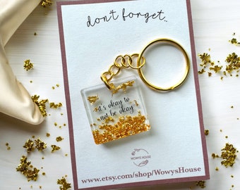 May include: Gold keychain with a clear square charm that says "it's okay to not be okay" and gold glitter inside. The keychain is attached to a gold key ring. The keychain is on a white surface with a pink card that says "don't forget..." and "www.etsy.com/shop/WowysHouse".