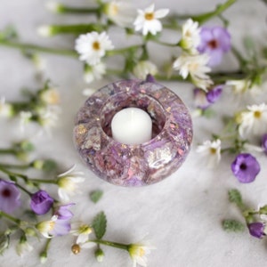 May include: A purple and gold candle holder with a white candle inside. The holder is made of resin and has a floral design. The holder is surrounded by white and purple flowers.