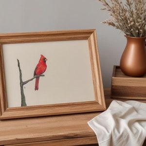 May include: A framed watercolor print of a red cardinal perched on a branch. The artwork is displayed in a light brown wooden frame. A copper-colored pitcher and a wooden box are in the background, with a white cloth draped on the table.