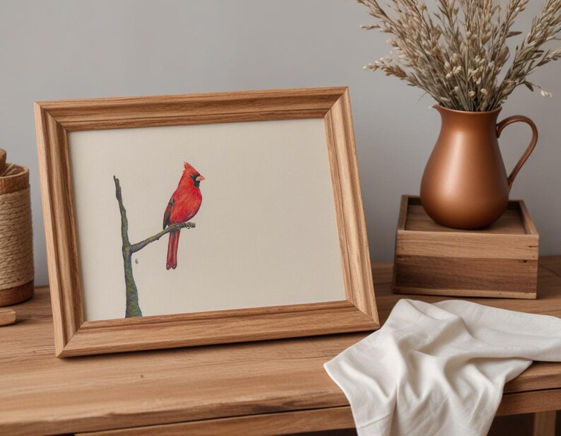 May include: A framed watercolor print of a red cardinal perched on a branch. The artwork is displayed in a light brown wooden frame. A copper-colored pitcher and a wooden box are in the background, with a white cloth draped on the table.