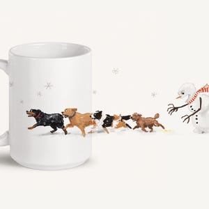 Op de afbeelding: Een witte keramische mok met een bedrukte illustratie van vijf honden die in een rij door een besneeuwd landschap rennen naar een sneeuwpop met een rood-wit gestreepte sjaal. Kleine sneeuwvlokken zijn verspreid rond de honden en de sneeuwpop.