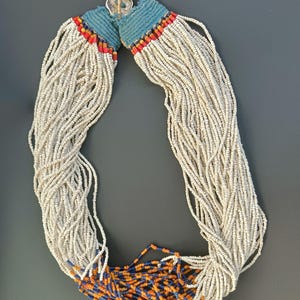 Könnte beinhalten: Eine lange weiße Perlenkette mit blauen und orangefarbenen Perlen am unteren Ende. Die Kette hat einen blauen Stoffquaste oben.