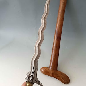 Pode incluir: Uma adaga de lâmina ondulada com acabamento metálico e cabo texturizado, acompanhada por uma bainha de madeira castanho-clara, lisa e separada. A lâmina apresenta uma borda sinuosa distinta e um guarda-mão metálico detalhado.