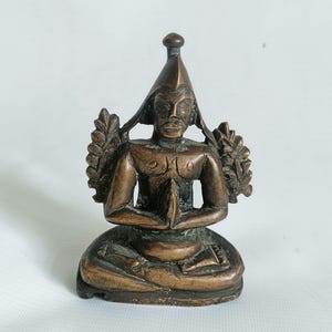 Puede incluir: Una pequeña estatua de bronce de una figura sentada con una corona y un tocado elaborado. La figura tiene una expresión serena y sostiene sus manos en posición de oración. La estatua está decorada con intrincados detalles y tiene una rica pátina.