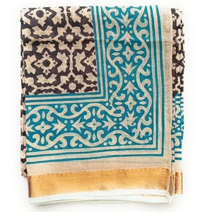 May include: A folded textile featuring a cream-coloured base with a dark brown geometric pattern on one side and a vibrant teal border with intricate scrollwork. The bottom edge is finished with a wide, metallic gold band with a textured weave.