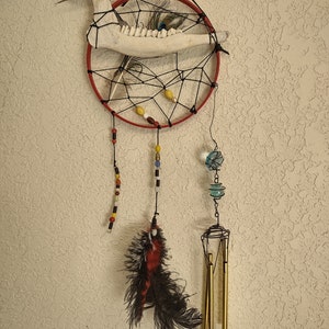 May include: A dream catcher with a red hoop, black netting, and a bone with peacock feathers. It has a variety of beads and a wind chime with gold tubes hanging from it.