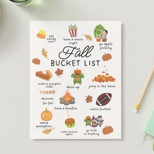 May include: A white Fall Bucket List print with illustrations of pumpkins, leaves, and activities like "eat candy corn" and "go trick or treating." A pencil and a small green notebook are on the right.