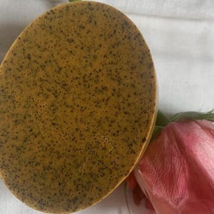 May include: Oval-shaped, golden-brown soap bar with dark speckles. A pink rose with green leaves is in the lower right corner. The soap has a textured appearance, suggesting natural ingredients.