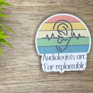 May include: A sticker with a rainbow background and a stylized ear with a heartbeat graphic. The text reads "Audiologists are Ear-replaceable."