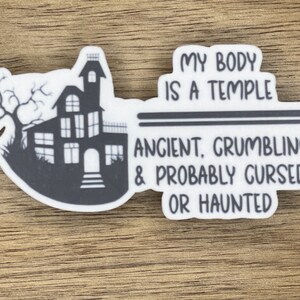 May include: A white sticker with a black silhouette of a haunted house and the text "My body is a temple - Ancient, Grumbling, & Probably Cursed or Haunted".