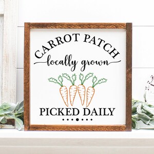 May include: A square wooden sign with a white background and a brown frame. The sign features the words "CARROT PATCH locally grown PICKED DAILY" in black script and a graphic of four orange carrots with green tops.