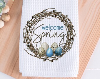 May include: White waffle-weave kitchen towel with a spring-themed design. The design features a wreath of twigs and pussy willows encircling the words "welcome Spring" and several speckled Easter eggs in shades of blue and beige.