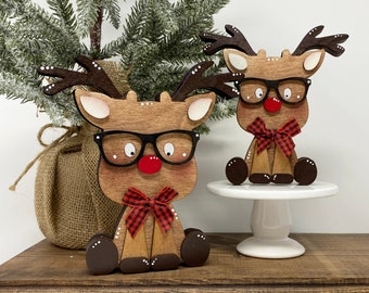 May include: Two wooden reindeer figurines with glasses and red noses. One sits on a white cake stand, the other on a wooden surface. Both have black-framed glasses, red and black plaid bows, and brown antlers. A small Christmas tree is in the background.
