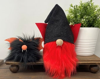 May include: Two Halloween gnome decorations. One is a black furry bat with orange wings. The other is a gnome with a black glittery hat, red beard, and red cape. They are displayed on a wooden tray.