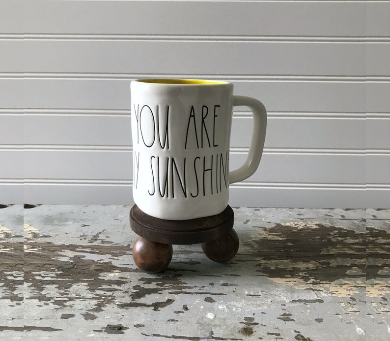 May include: White ceramic mug with the text "YOU ARE MY SUNSHINE" in black lettering. The mug has a yellow interior and sits on a dark brown, three-legged wooden stand. The background is a white wall.