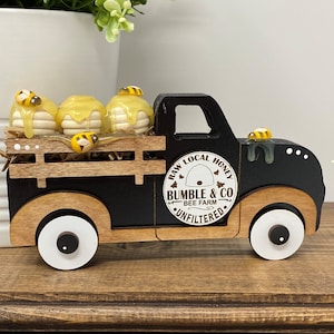 May include: A black wooden toy truck with a honey theme. The truck bed carries three honeycombs with dripping honey and bee figurines. The truck features a "Bumble & Co" logo and the words "Raw Local Honey" and "Unfiltered".