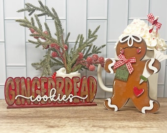 May include: A wooden gingerbread man decoration with a red and white gingham bow and a tag that says "Christmas calories don't count." A sign that says "Gingerbread cookies" is in front of a mug with a sprig of berries.