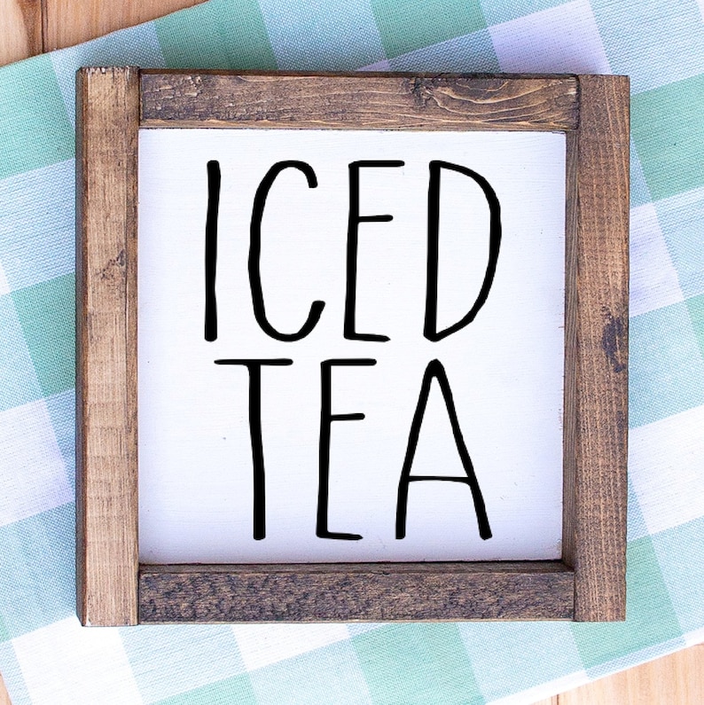 May include: A square wooden sign with the words "ICED TEA" in large, black, handwritten-style letters. The sign has a white background and is framed with dark brown wood. The sign is placed on a green and white checkered cloth.