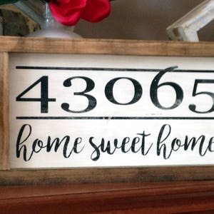 May include: Wooden framed sign with the numbers "43065" and the phrase "home sweet home" in black lettering on a white background. The frame is a light brown color.