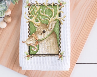 May include: White waffle-weave kitchen towel featuring a detailed illustration of a reindeer with golden antlers and a green and red harness. The reindeer is set against a green background with falling snow, framed by a black and white checkered border and holly accents.