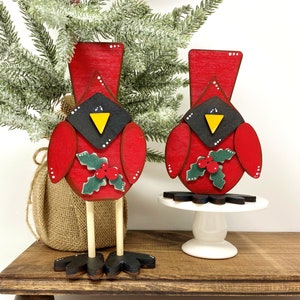 May include: Two wooden cardinal figurines. The cardinals are painted red with black and yellow accents. Each has a holly berry design. One stands on wooden legs, the other on a white pedestal. A Christmas tree is in the background.