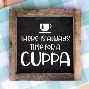 May include: Square wooden sign with a black background and white text that reads "THERE IS ALWAYS TIME FOR A CUPPA". The sign has a white graphic of a teacup and a tea bag. The frame is made of brown wood.